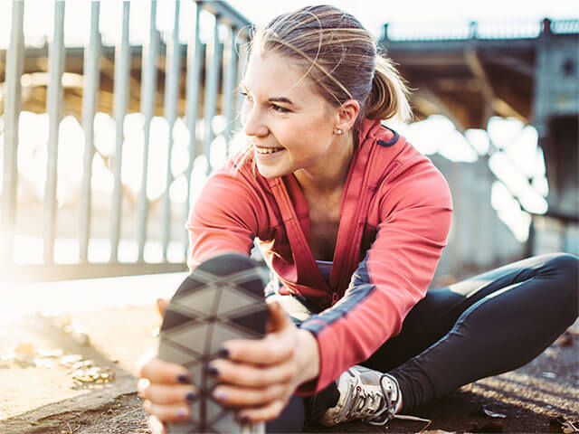 Ung kvinne i treningsklær tøyer ut utendørs, smiler mot noe utenfor bildet.